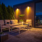 An outdoor patio at night with cushioned chairs positioned around a table. Downlights shine on this display. Next to the patio is a carport with a car visible through the glass door.